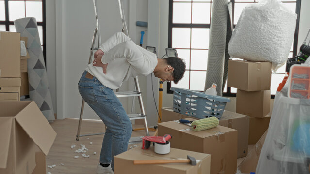 Man clutching lower back while bending over packed cardboard boxes and a ladder amid moving supplies inside a building; pain backache strain.