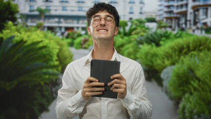 Man holding tablet with hands visible, smiling and looking up on a street lined with green bushes and buildings  quiet joy. © Krakenimages.com