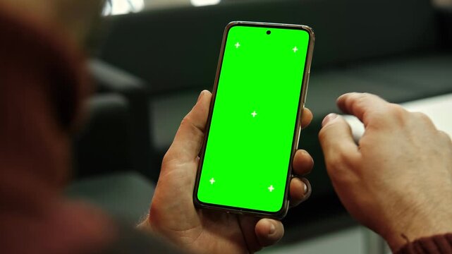 Hand of young man holding smartphone green screen in business office work. Mobile phone vertical mockup. Male person using phone with blank green background display. Chroma key smart phone. Close up.
