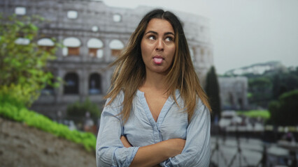 Fototapeta premium Woman sticks tongue out and makes a mocking hand gesture with folded arms in front of coliseum building on an outdoor plaza; playful travel humor.