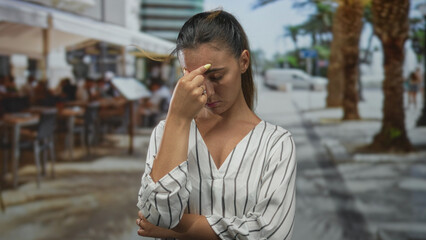 Fototapeta premium Woman pinching nose and pressing forehead at a restaurant terrace with blurred diners and palm trees in street view; stress exhaustion.