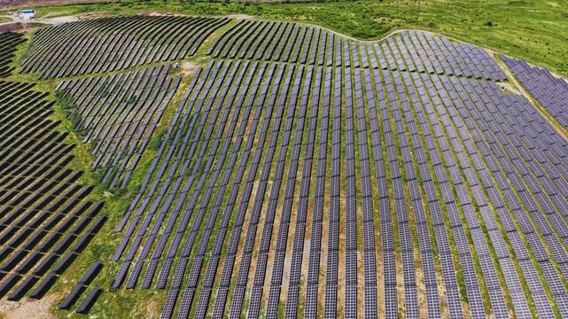 Solar panels stretch across a large area of land under clear skies. The field is organized in rows and is surrounded by green fields and patches of grass.
