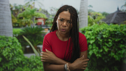 Fototapeta premium Woman with hands to mouth and arms crossed in park near palm tree, wearing red top and bracelets; discomfort cold.
