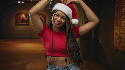 Naklejka premium Woman adjusts santa hat with raised arms, smiling and showing bare midriff in an art gallery building lit by warm paintings and soft spotlights; festive joy.