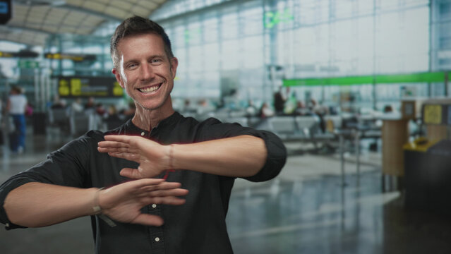 Man with arms outstretched showing forearms and hands while smiling in busy airport terminal with seating and checkin area visible; happiness welcome.