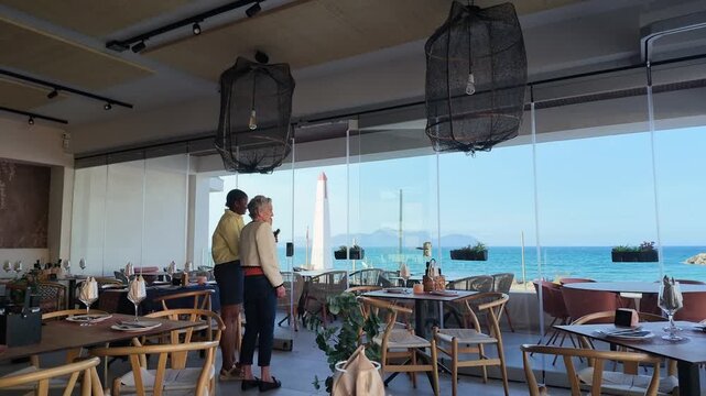 Can Picafort Mallorca Spain. 01.06.2026. Video. Modern dining room with panoramic windows overlooking the turquoise sea and coastline, featuring stylish tables and patrons select a place to sit.