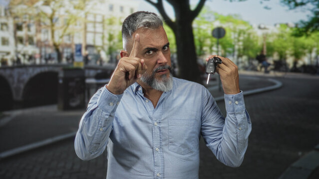 Man holding car keys in his raised right hand and points finger upward with left hand on a city street by a canal, wearing light blue shirt and gray beard; warning.