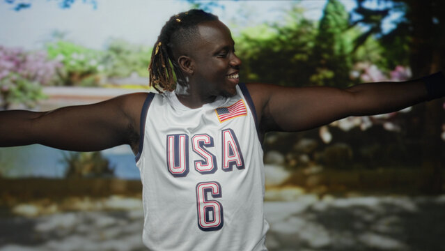 Young african american man in white usa basketball jersey with flag patch, arms outstretched and smiling in a green park with trees and sunlight; joy celebration.