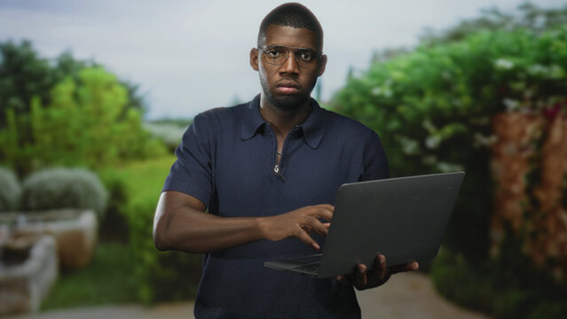 Man holding laptop and typing with right hand while looking at screen in studio setting with blurred plants; focused productivity.