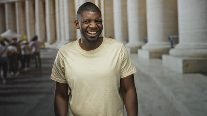 Obraz premium Man squints and smiles showing teeth while standing among stone columns in a public building with blurred pedestrians; uncertainty introspection.