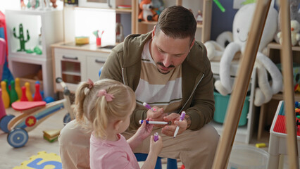 Obraz premium Man teaching girl, handing purple marker and guiding her hand at a play easel amid toys and books in building; tenderness learning.