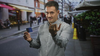 Naklejka premium Man holding credit card beckons with open hand on street cafe sidewalk, moustache and checkered shirt visible; confident transaction.