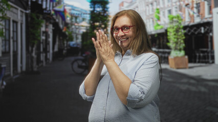 Fototapeta premium Woman in a light striped shirt and red glasses pressing her hands together in a thankful clap while smiling on a narrow city street lined with outdoor cafes; gratitude joy.