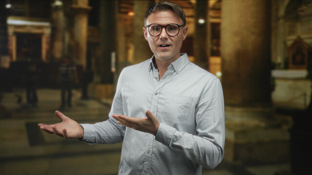Man with glasses and white shirt gesturing open hands and speaking in a cathedral interior with marble columns and altar; reflection.