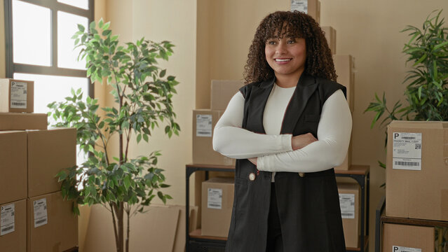Woman with cardboard boxes and parcel labels, arms crossed and smiling in a building delivery office, surrounded by stacked boxes and plants; confidence entrepreneurship.