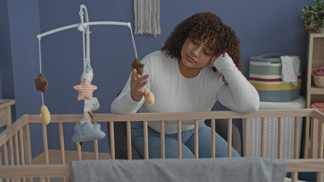 Woman leaning on crib with hand to head, touching mobile and peering into empty cradle in building; exhaustion.