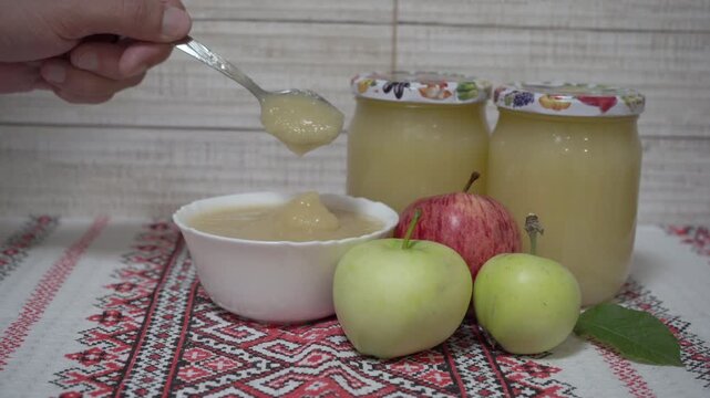 Glass jars of applesauce and fresh apples, tasting apple jam from a spoon, close-up.