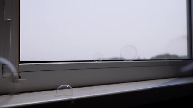 Glistening, vibrant soap bubbles gracefully drift downwards, landing and popping on a sunlit interior windowsill. This lighthearted visual is perfect for themes of happiness and gentle