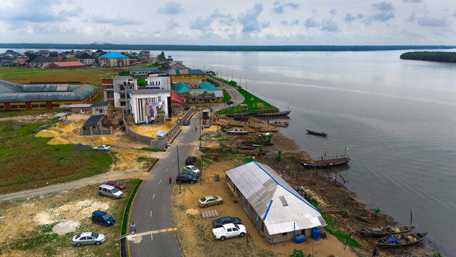 Aerial view of the coastal settlement with buildings, a road, parked cars, and wooden boats moored along the riverbank in Minima, Rivers, Nigeria.