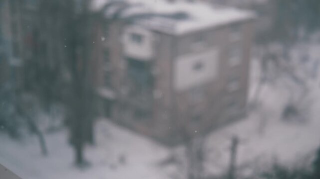 Snowflakes fall slowly against a defocused background of a brick apartment building and bare trees. Static shot with shallow depth of field captures a melancholic and cold atmosphere.