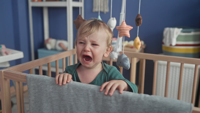 Boy crying with hands gripping crib rail and gray blanket over rail in building nursery, mobile toys above; distress need comfort.
