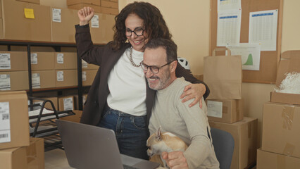 Fototapeta premium Man holding small dog and high fives woman at laptop amid parcel boxes in packing building; teamwork joy entrepreneurship.