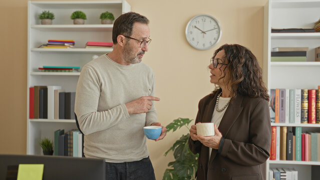 Man holding blue mug and gesturing with finger while talking to woman near bookshelf and wall clock in office; friendly chat.