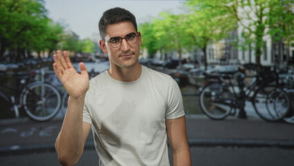 Naklejka premium Man waves hand on a canal street in amsterdam wearing glasses and a white t shirt; friendly greeting.