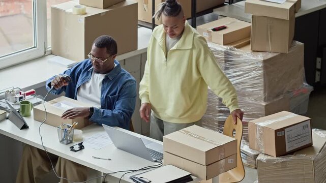 High angle view of African American man scanning barcode on box while multiethnic woman works on laptop in warehouse. Video represents e-commerce, logistics, and small business shipping operations