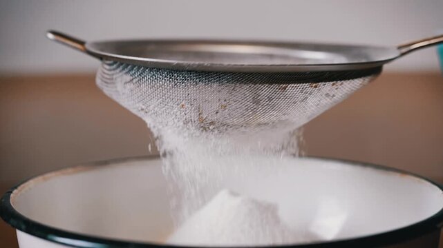 White flour is being passed through a fine metal sieve into a large white ceramic mixing bowl. Concept of home baking, culinary arts, precision, and healthy homemade cooking.