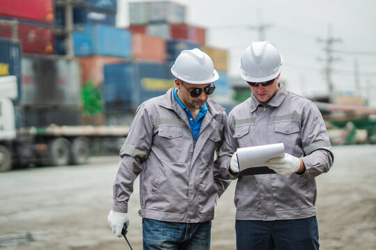 Logistics workers examine shipment paperwork and verify transportation details in container yard, ensuring cargo inspection accuracy and delivery coordination.