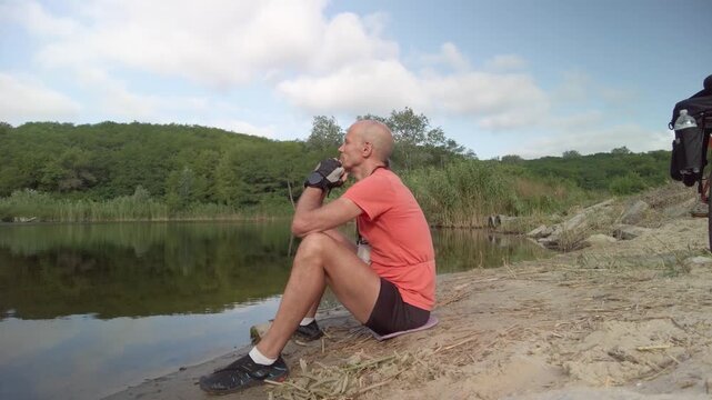 Male cyclist sitting on ground near lake taking break to rest Bicycle traveler relaxing near water and reflecting during journey pause in outdoor environment