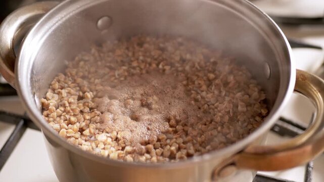 Buckwheat groats boiling in a stainless steel pot on a gas stove burner with visible steam and bubbling water. This cooking scene represents the concept of preparing healthy whole grains.
