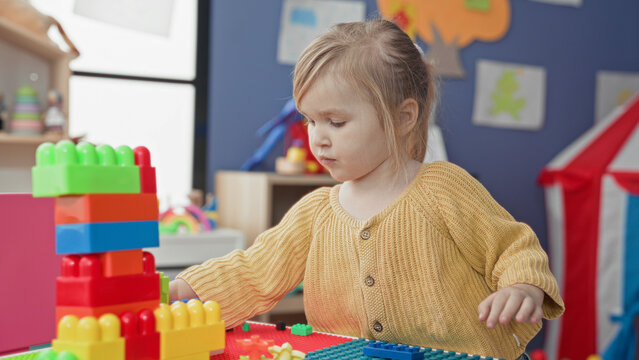 Girl in yellow sweater stacking colorful plastic blocks at a play table in a children's playroom inside a building; quiet concentration.