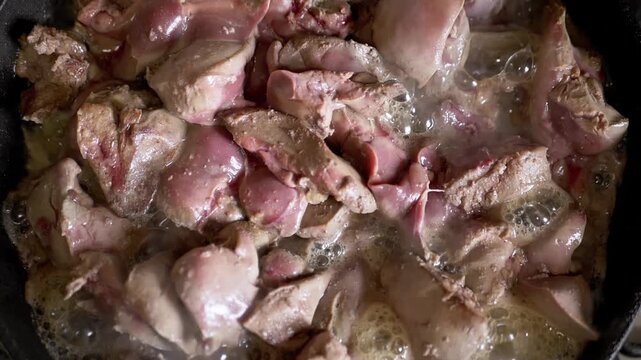 Raw chicken liver searing in a hot metal frying pan with bubbling oil. Culinary preparation of offal in a kitchen environment is the primary concept represented in this visual.