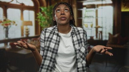 Obraz premium Woman in plaid shirt and white tshirt shrugs with hands up in a room, braids and nosepiercing visible, palms open and gaze upward; confusion.