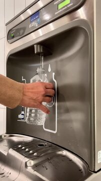 A person holds a water bottle under a refill station to fill it with water in a bright indoor space.