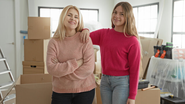 Mother and daughter smile, daughter has arm around mother's shoulder amid moving boxes in building; family warmth.