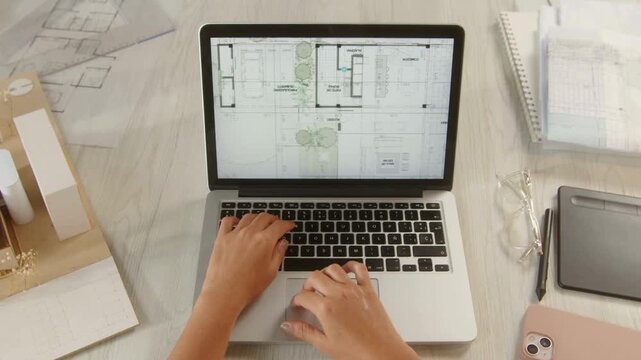 Architectural floor plan on laptop screen, architect hands typing on keyboard, professional design workspace with blueprints and scale models
