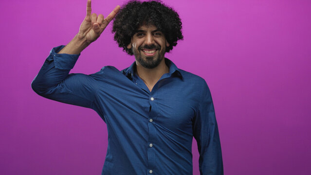 Curly haired man making rock horns gesture with raised hand and smiling in magenta studio; playful fun energy.