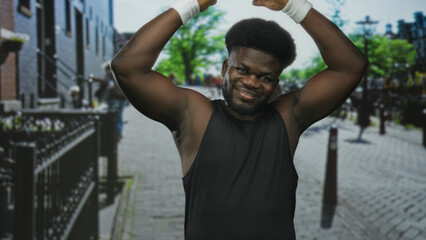 Fototapeta premium Young african american man raising bare arms overhead on city street in black tank top with white wristbands and smile; confidence fitness motion.