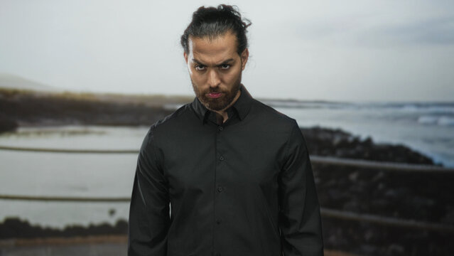 Man, young arab hispanic with beard and tied hair, in black shirt furrows brow and looks down at seaside railing on rocky coast; brooding solitude.