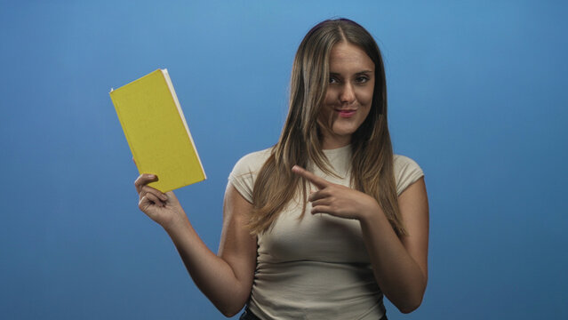 Woman holding a yellow book points finger to the cover in a studio, smiling while presenting the hardcover; confidence learning inspiration.