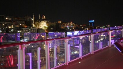 Obraz premium Night rooftop terrace glass balustrade in sharp foreground with blurred urban skyline and colorful bokeh lights, defocused night view city; background backdrop copyspace calm.