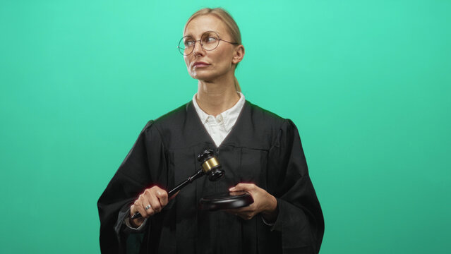 Woman judge in black robe raises gavel with right hand over wooden sounding block in studio green screen, wearing glasses and white collar; authority justice.