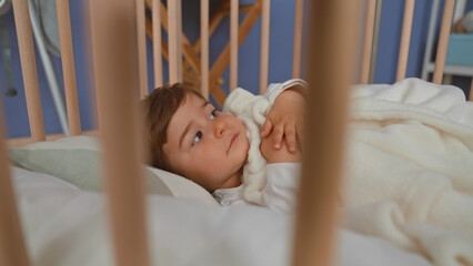 Boy lying in pale wooden crib clutching soft fleece blanket, hand near mouth, gaze sideways toward room window in building  cozy calm. © Krakenimages.com