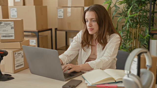 Woman types on laptop while handling stacked parcels and a scanner in a small business building; focused entrepreneurship.