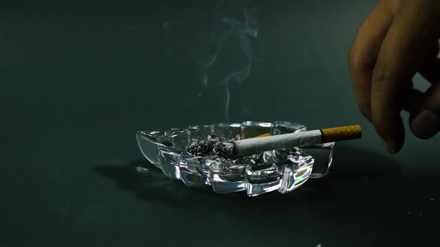 Hand holding a lit cigarette over a glass ashtray with smoke rising tobacco nicotine smoking addiction finger