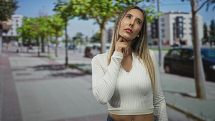 Fototapeta premium Woman with exposed navel piercing and hand on chin on street, wearing white ribbed top and denim jeans, looking upward and tilting head; contemplation focus.