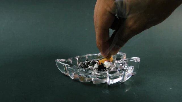 Hand placing a lit cigarette into a clear glass ashtray with ash and smoke tobacco nicotine smoking addiction habit